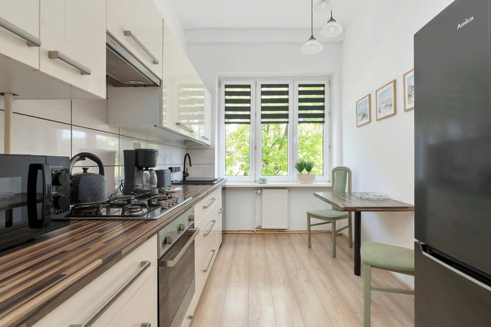 A kitchen corridor extends toward windows at the far end with cabinets and appliances arranged along the walls. Wood textures contrast with white finishes.