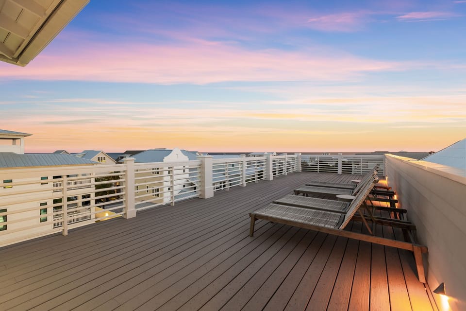 A rooftop terrace overlooks neighboring properties at sunset, featuring lounge chairs on dark wood decking and white railings. Soft accent lighting lines the deck's perimeter, creating a peaceful outdoor setting.