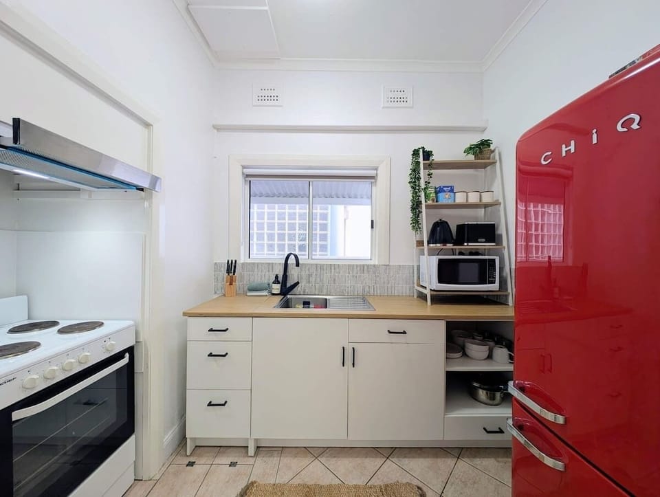 Enjoy preparing meals in this cheerful kitchen, featuring white cabinetry, a wooden countertop, a black gooseneck faucet, an open appliance shelving unit, and a bold red retro refrigerator that completes the space with character.