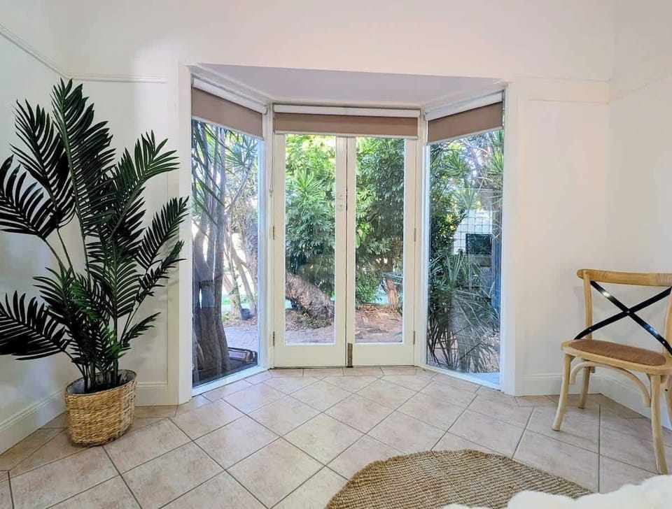 Step into this sunlit nook featuring French doors, floor-to-ceiling glass bay windows overlooking garden greenery, tiled flooring, a potted palm plant, and a classic wooden cross-back chair.
