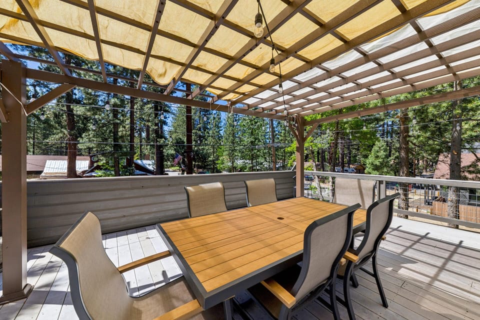 Covered deck with a wood dining table, six beige sling chairs, string lights, and views of tall pine trees.