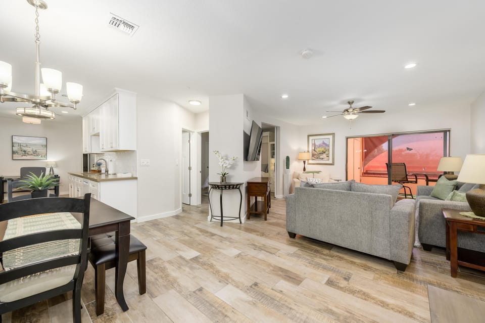 Open-concept living and dining area with wood-look tile flooring, gray couches, ceiling fan, and sliding balcony doors.