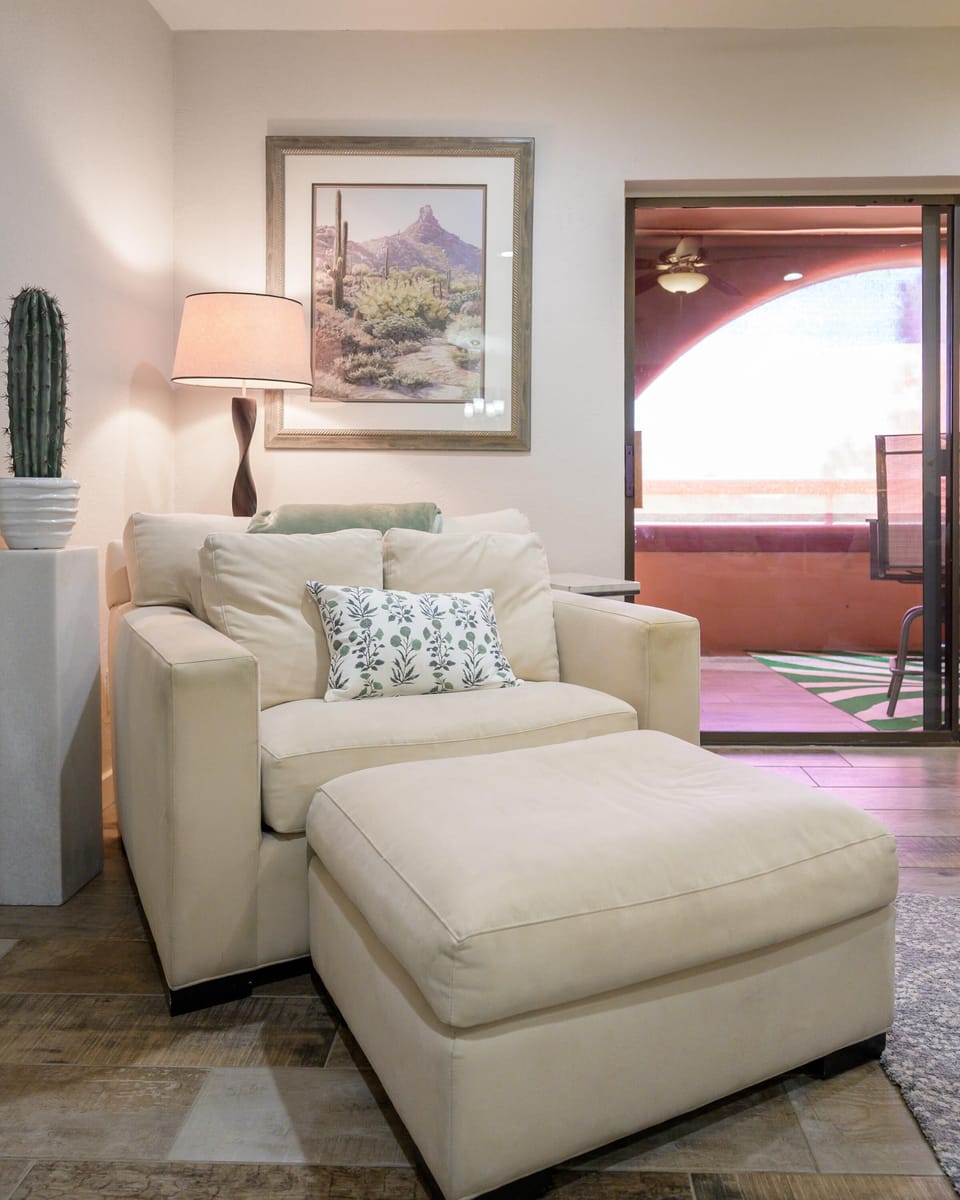 Cream armchair and ottoman sit on wood-look tile flooring beneath a desert landscape print and standing lamp.