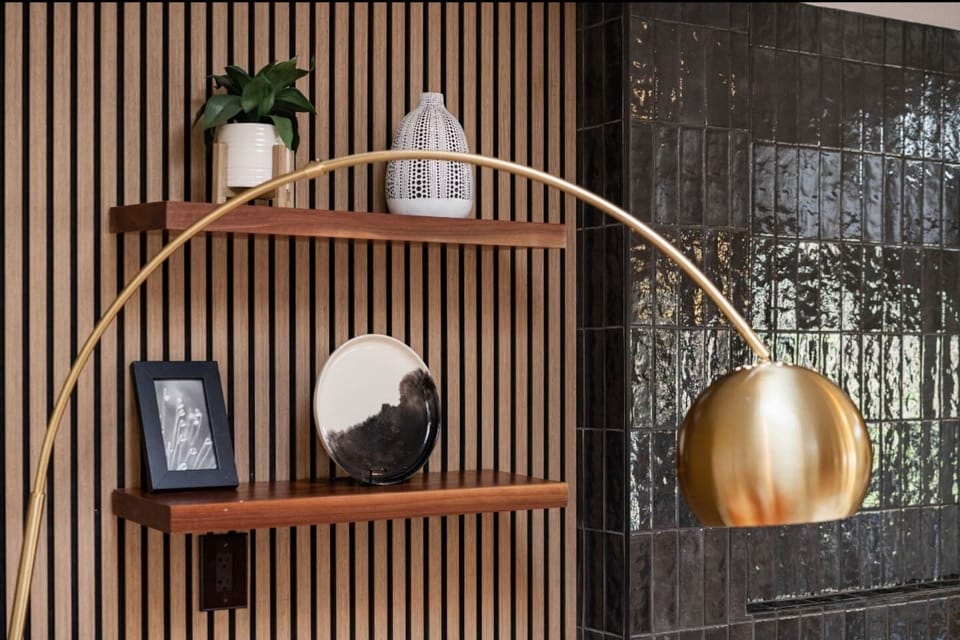 A mid-century brass arc lamp casts a warm glow over wood shelves against walnut acoustic panels. Styled with ceramics, art, and greenery, the space blends warmth and modern elegance.
