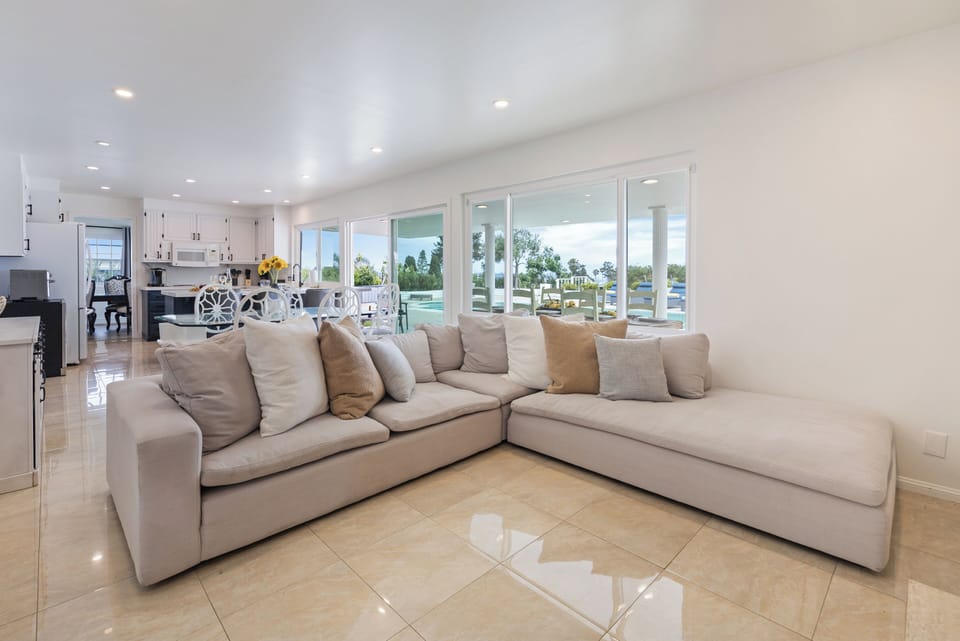 Bright living room with plush seating, a fireplace, TV, and poolside views through oversized windows.