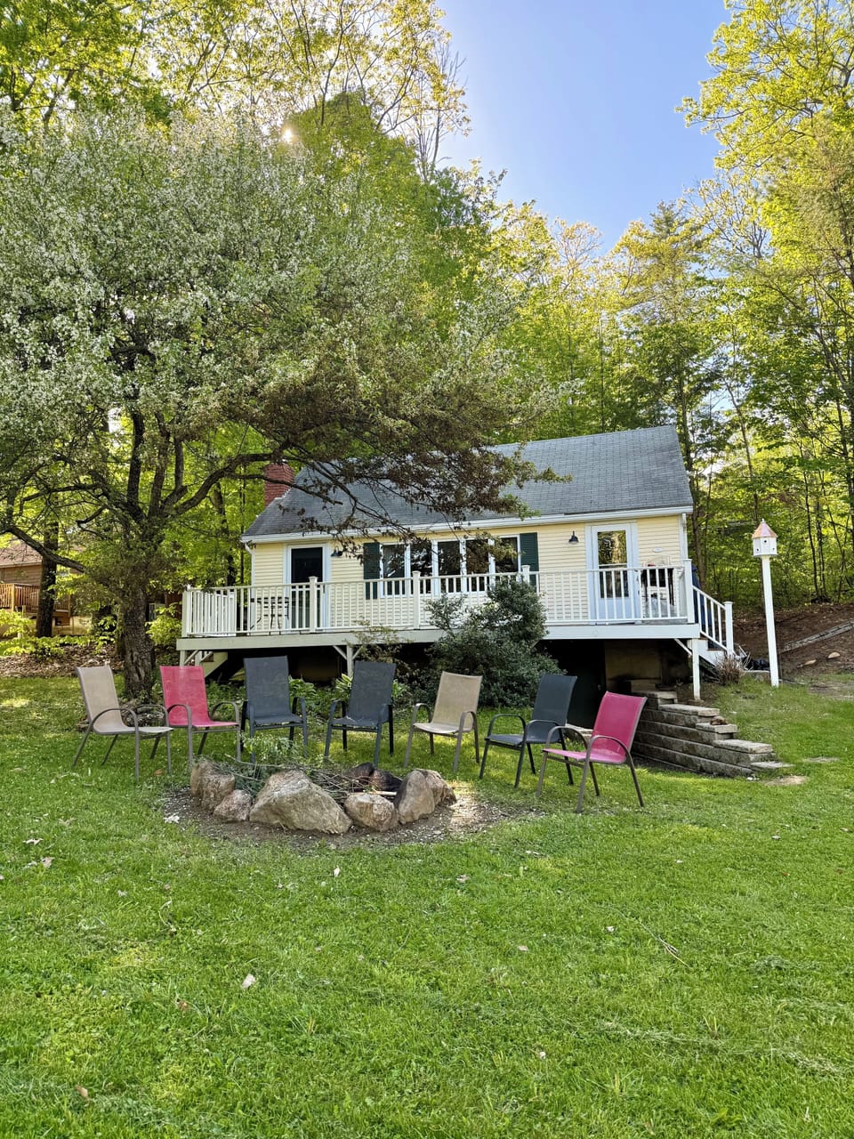A charming cottage nestled in a wooded setting with lush green lawn, outdoor seating around a fire pit, and a welcoming front deck.