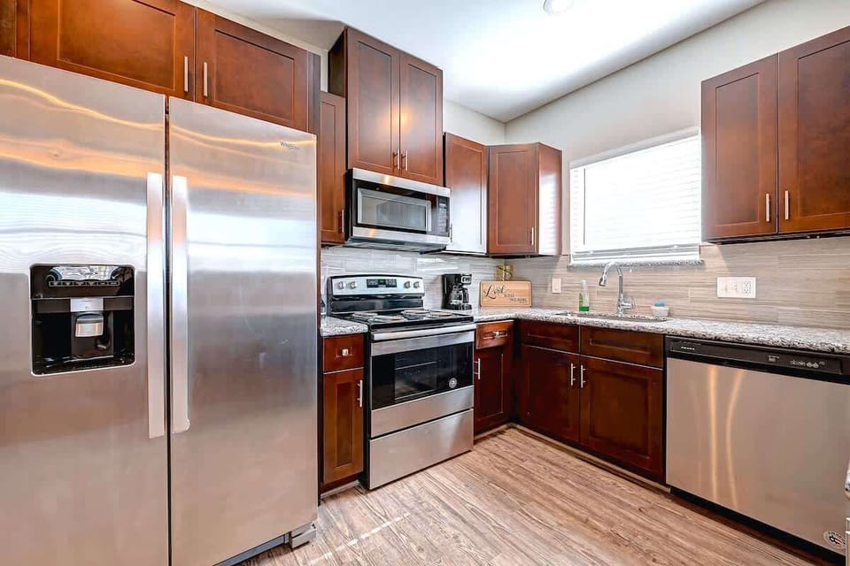 Modern kitchen with stainless steel appliances, granite countertops, and rich wood cabinetry. Features a built-in microwave, dishwasher, and double-door fridge, with ample natural light from a large window and stylish tile backsplash.