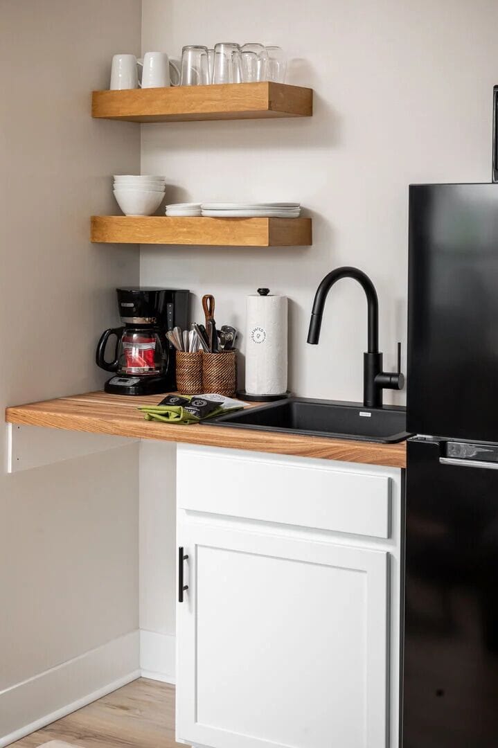Compact kitchenette with open shelving, dishware, coffee maker, sink, and full-size fridge for easy in-room convenience.