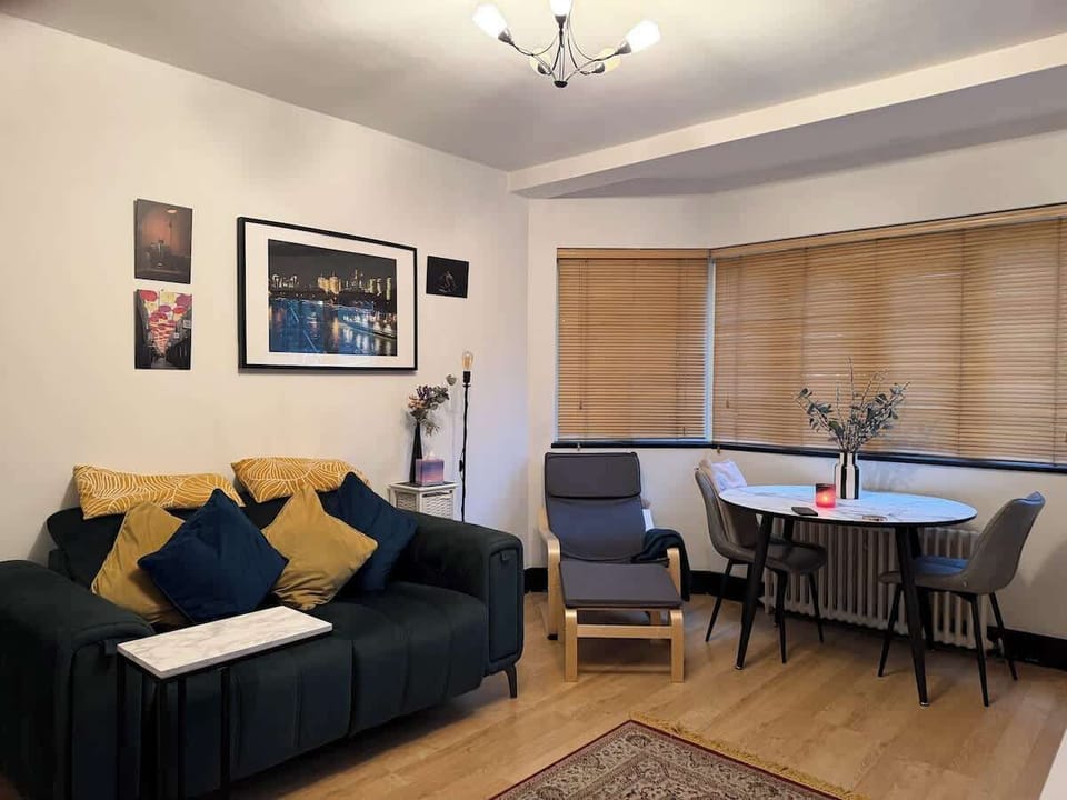 A cozy living room features a dark green sofa with yellow and blue pillows, an armchair, and a small dining table set near a large window with wooden blinds.