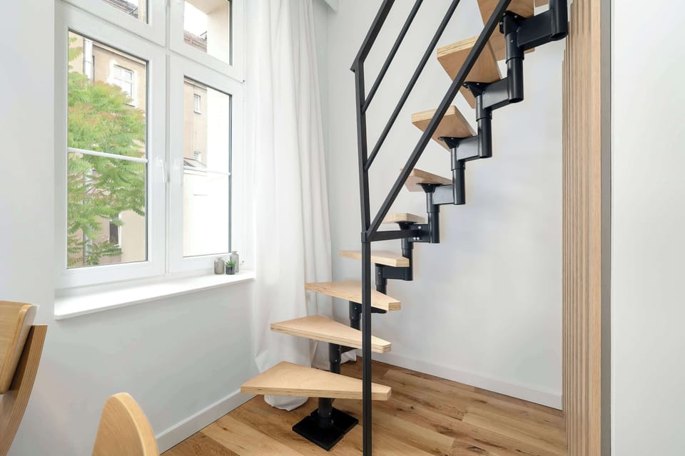 A staircase with wooden steps rises beside large windows that illuminate the space. The design combines modern metal framing with warm materials.