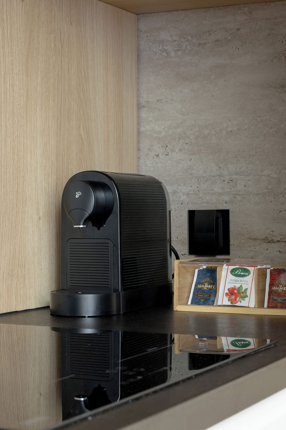 A sleek coffee station features a matte black capsule espresso machine sitting on a glossy black countertop. A small container with coffee pods sits neatly beside the appliance against a textured backsplash.