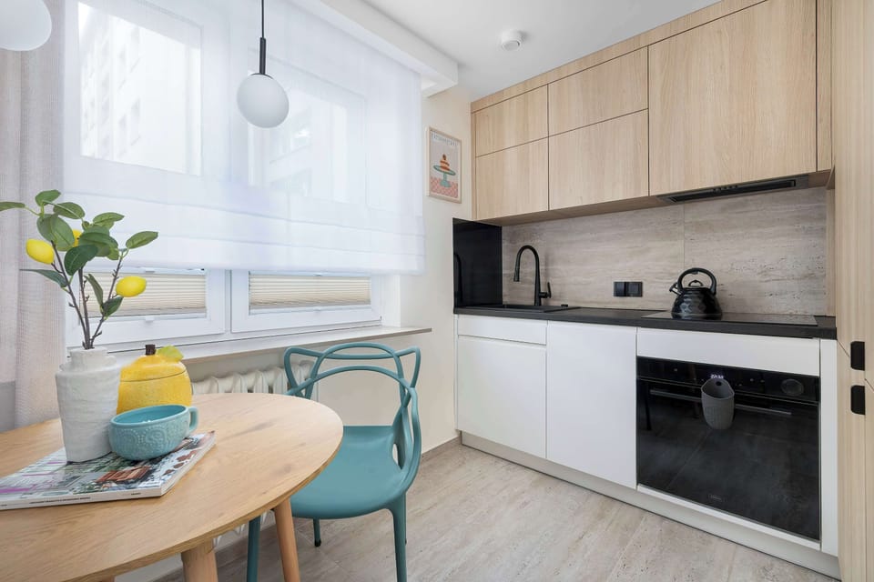 A bright kitchen blends white lower cabinets with warm wooden upper cabinetry and features a round dining table with a turquoise chair. Modern built-in appliances, including an oven and a black induction hob, complete the functional space.