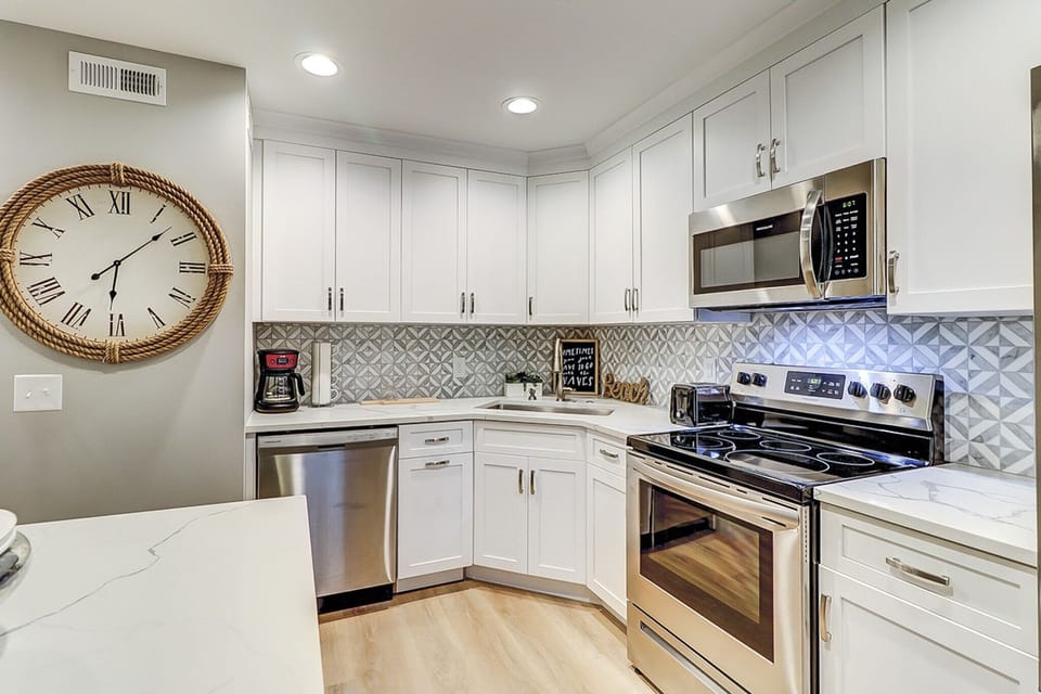 Alabaster cabinetry and a geometric backsplash create a bright, contemporary culinary space. Stainless steel appliances gleam against marble counters, while the oversized rope clock adds a charming nautical touch to this airy kitchen.