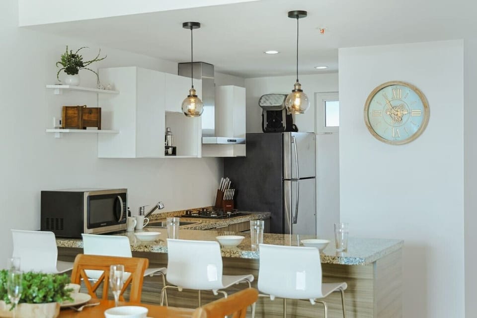 A clean, contemporary open-concept kitchen featuring white cabinetry and granite countertops. The space is equipped with modern stainless steel appliances—including a refrigerator, stove, and microwave—and features a breakfast bar lined with white chairs