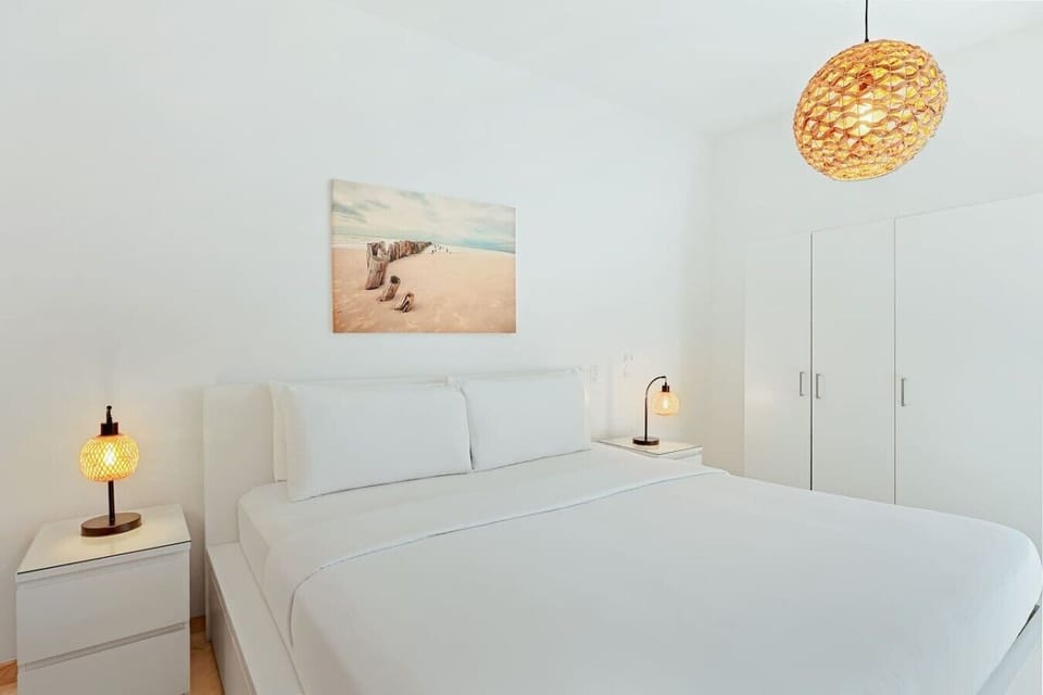 An alternative view of the bedroom area, showing the king-sized bed flanked by two white nightstands with matching woven bedside lamps. A scenic beach art piece hangs on the wall above the headboard, and a white closet is situated to the right.