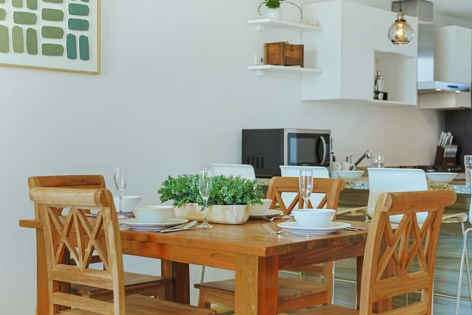 A close-up view of a wooden dining table set for a meal, decorated with a green planter box as a centerpiece. The open-concept background gives a glimpse of the kitchen area, complete with white cabinets and a microwave.