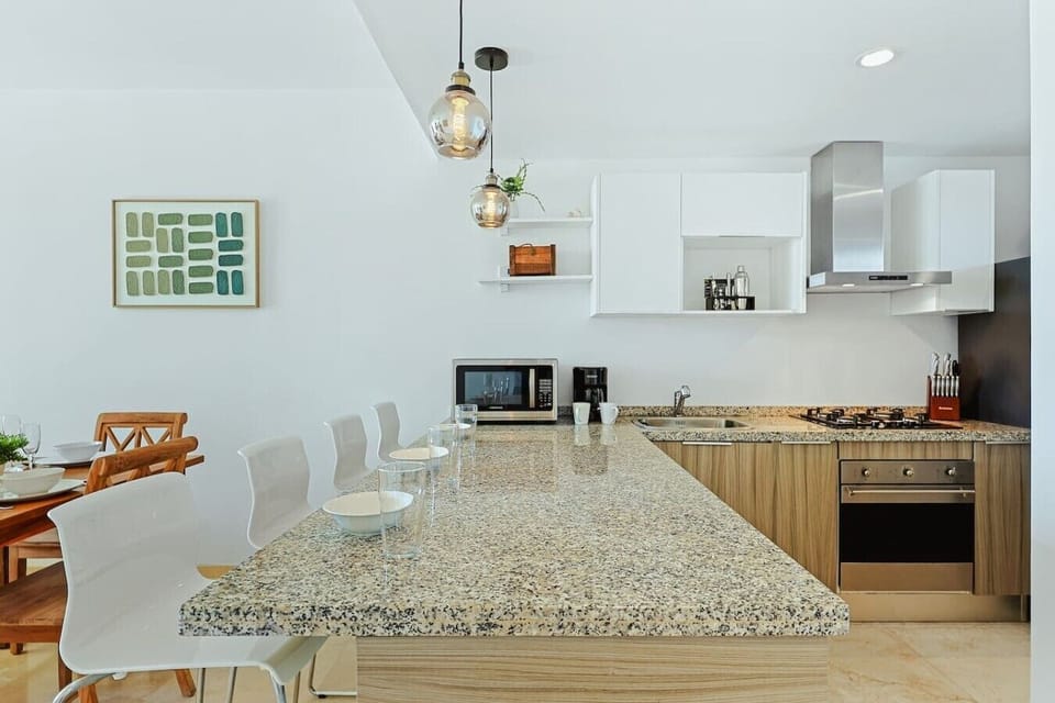 A front-facing view of the extensive granite breakfast bar island, lined with white modern stools and illuminated by low-hanging glass pendant lights. The background shows the kitchen sink, a countertop microwave, and a small green art piece on the left w