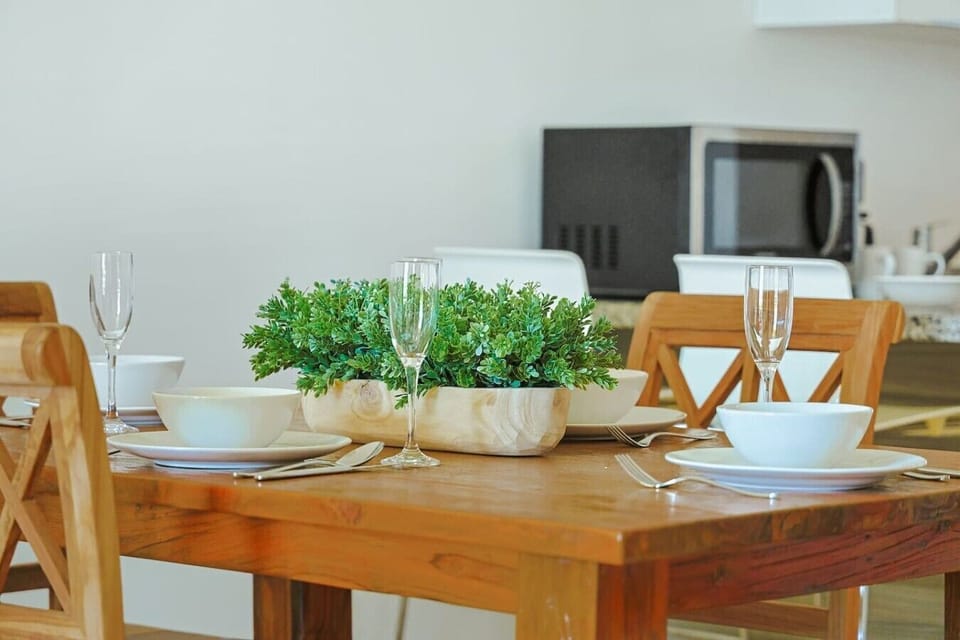 A close-up focus on the wooden dining table, nicely set with white ceramic plates, matching bowls, and elegant champagne flutes. A simple wooden box planter filled with green foliage acts as the table`s centerpiece.