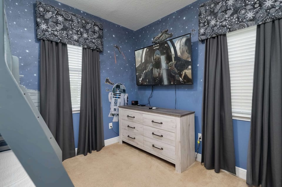 Star wars-themed room with a wall-mounted television and dresser between two windows with dark curtains.