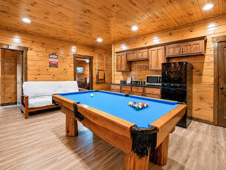 Game room with a blue-felt pool table, sleeper sofa, refrigerator, microwave, wood cabinetry, and open floor space for relaxing between games.