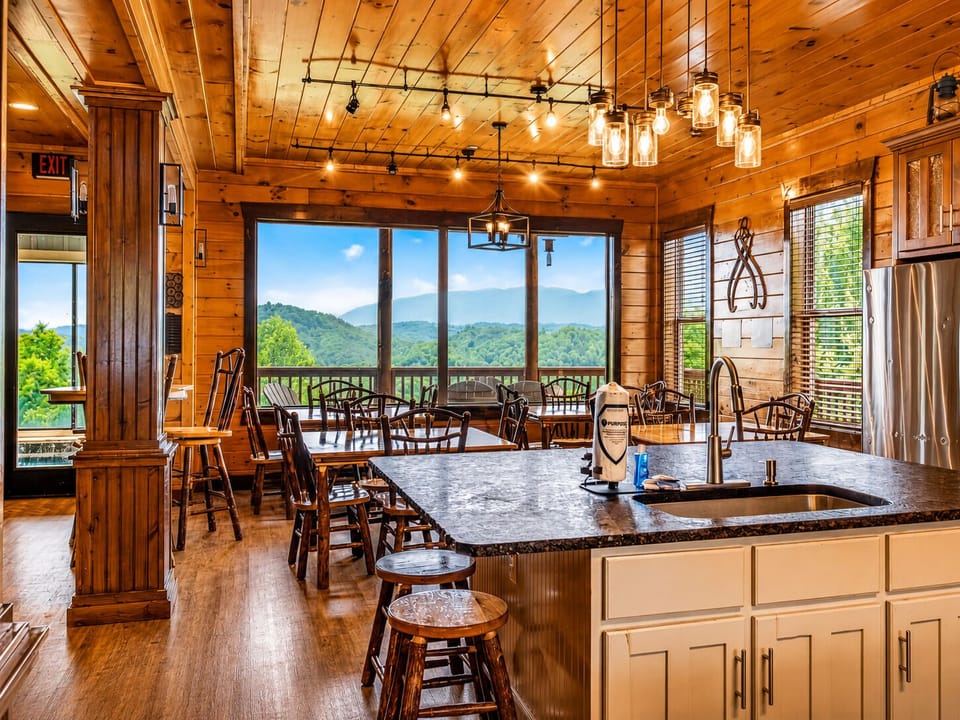 Open kitchen and dining area with granite island seating, pendant lighting, wood cabinetry, stainless-steel appliances, and large windows overlooking the Smoky Mountains.