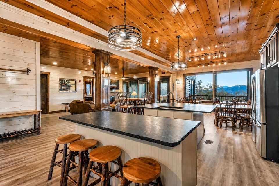 Fully equipped cabin kitchen with a large granite island, barstool seating, pendant lighting, rustic wood ceiling, and open access to the dining and living areas.