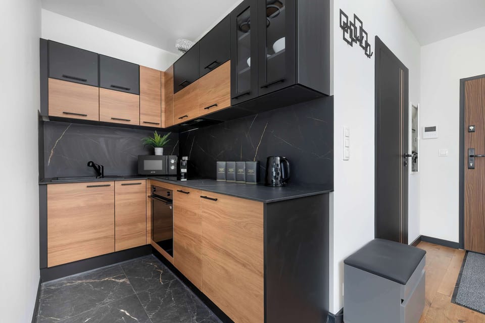 A wider angle of the kitchen area displays the seamless integration of wood and black cabinetry stretching toward the entryway. A simple dark trash bin sits at the edge of the kitchen zone.