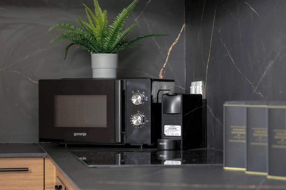 A close-up shot focuses on a black microwave and a coffee machine placed on the kitchen counter against the dark marble tile background. A small green plant adds a touch of color to the corner.