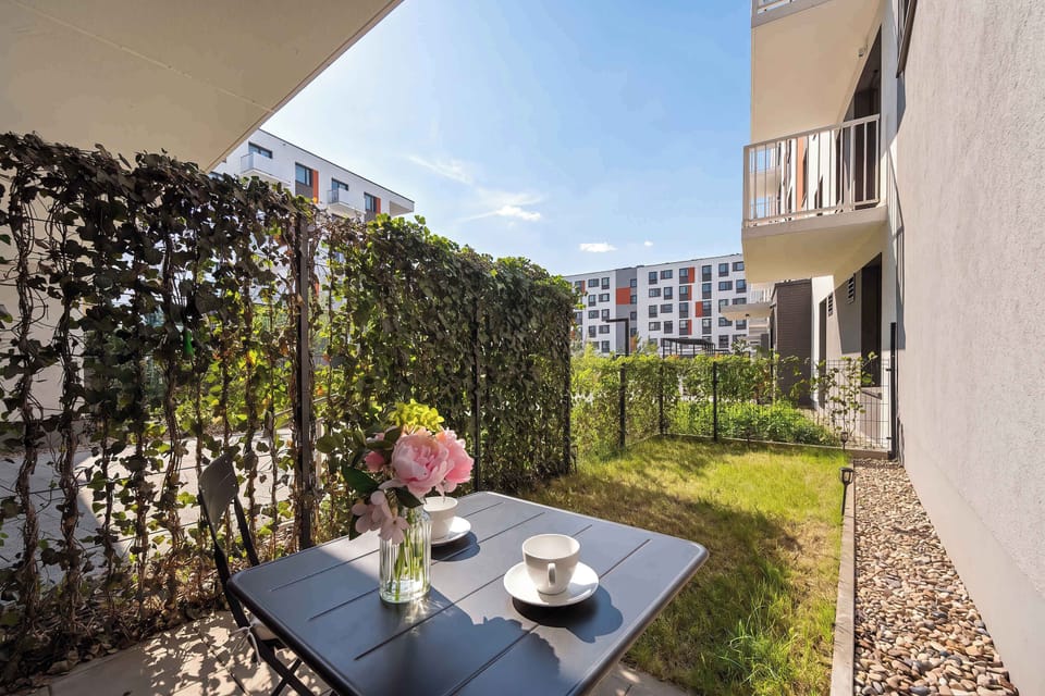 A small outdoor dining set consisting of a black table and chairs sits on a paved patio right outside the apartment. A dense green hedge creates a private oasis separate from the neighboring buildings.