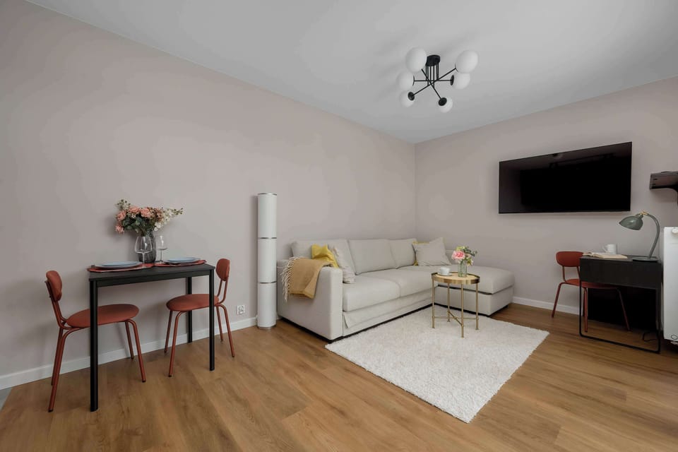 The spacious living room is shown from a corner perspective, highlighting a small dining table with red chairs on the left. A comfortable sofa and a study desk occupy the main living area.