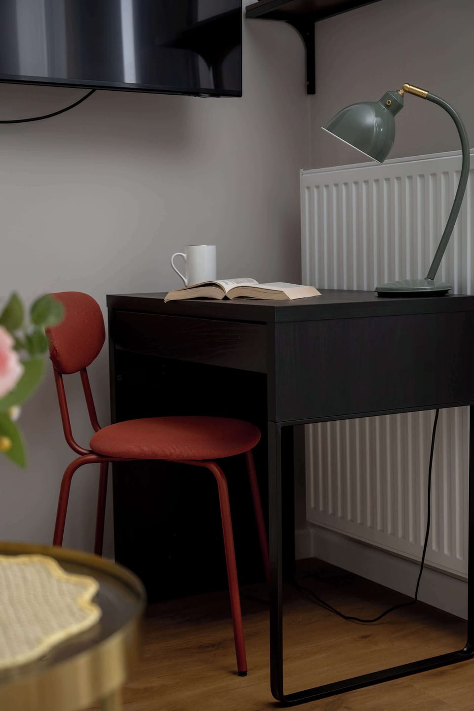 A close-up shot focuses on a compact black study desk paired with a stylish red chair. A small desk lamp provides targeted lighting next to a sleek wall-mounted television screen.