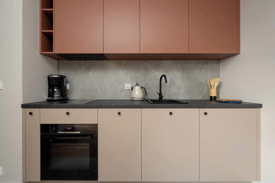 The sleek kitchen area displays minimalist cabinetry with a muted peach upper section and beige lower cabinets. A built-in oven, a black sink, and a modern faucet accent the clean gray backsplash.