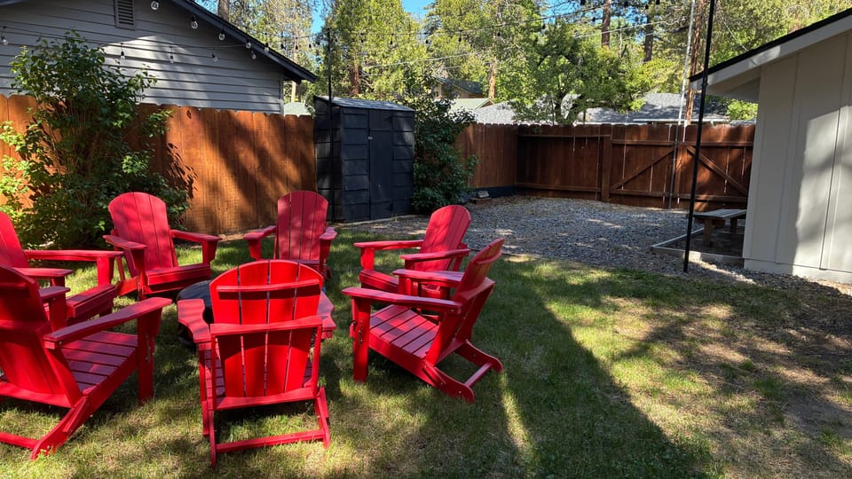 Fenced backyard with string lights, red Adirondack chairs, picnic table, shed, and pine trees.