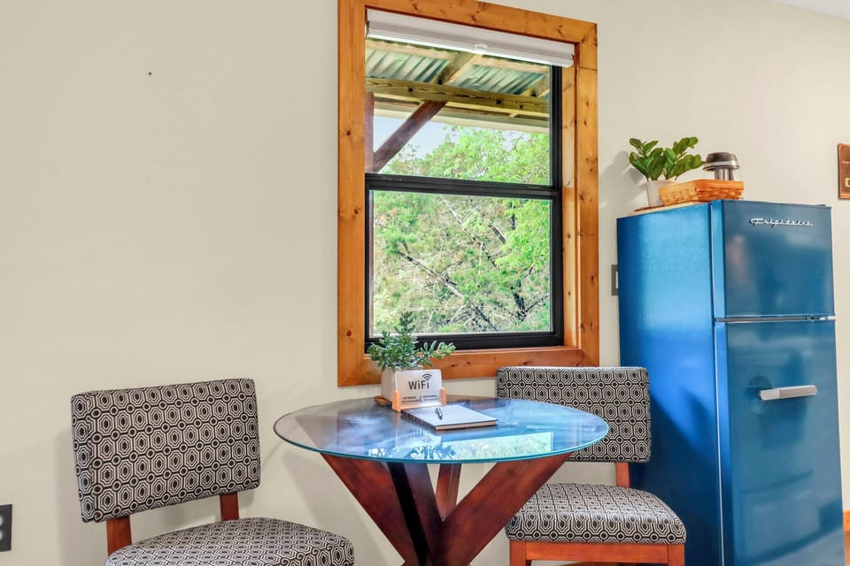 Your cozy dining nook features a charming bistro table with modern chairs and a retro blue fridge, all framed by peaceful garden views through rustic wooden windows.