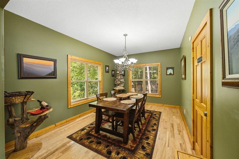Picture yourself savoring meals at this handsome wooden table, bathed in natural light from large windows that frame peaceful greenery beyond. The cozy chandelier and warm wood floors create a welcoming space.