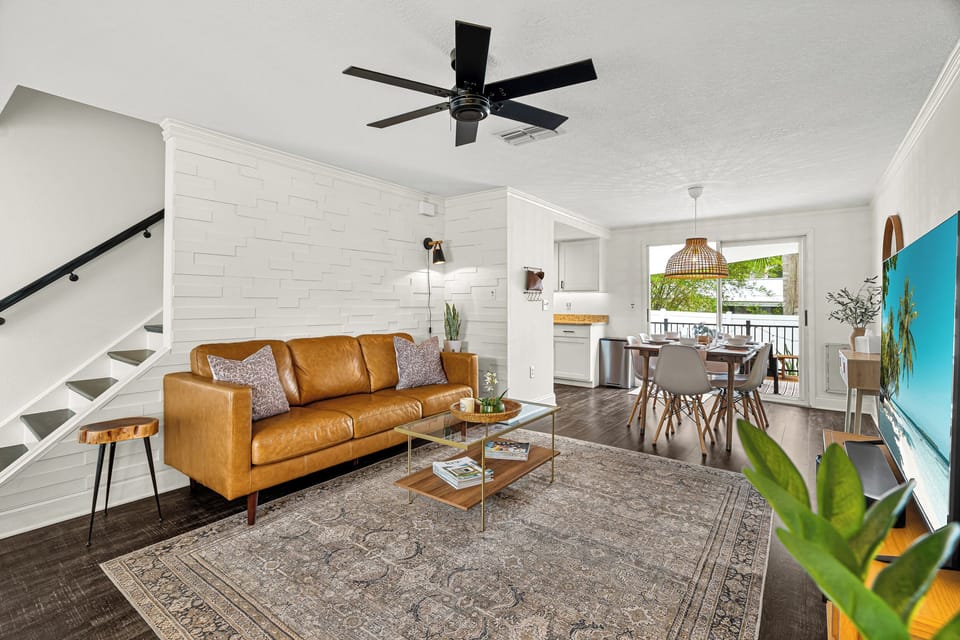 Picture yourself relaxing on the caramel leather sofa, surrounded by bright coastal decor and natural light streaming through the windows overlooking a peaceful outdoor view. The open layout invites you to unwind effortlessly.