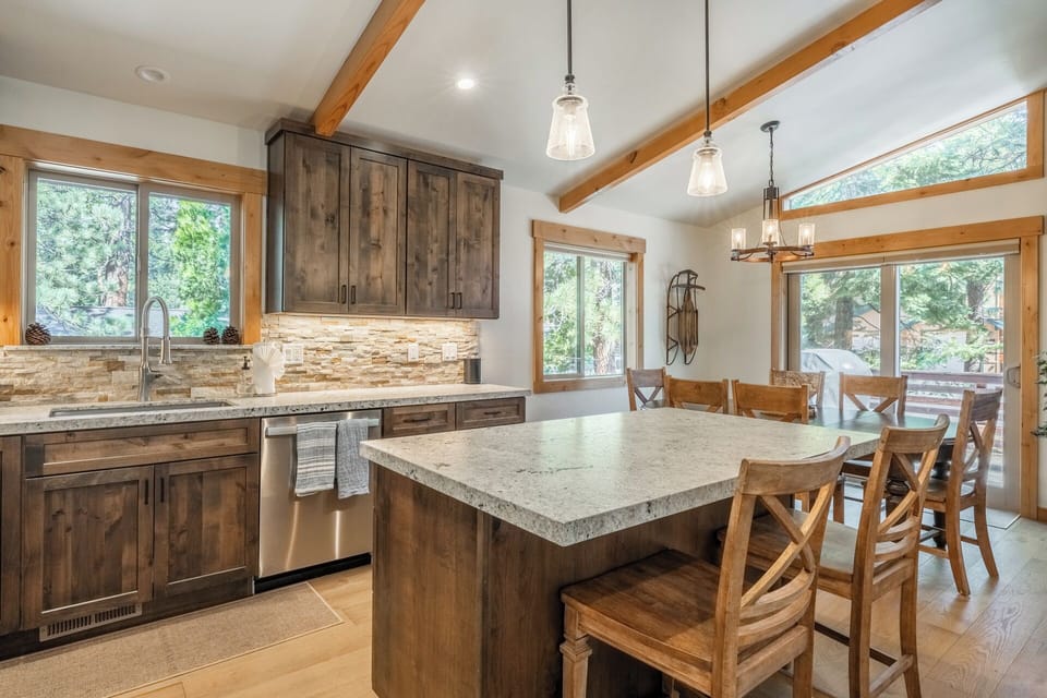 A cabin-style kitchen and dining area featuring rustic wood cabinetry, exposed beams, and modern granite countertops. Natural light streams through multiple windows overlooking surrounding pine trees, creating an inviting mountain retreat atmosphere.