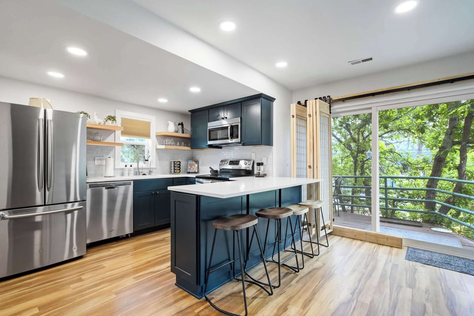 The fully stocked kitchen is designed to be used, with professional-grade stainless appliances, a French door refrigerator, open wood shelving, and large windows that frame the treetops while you cook.