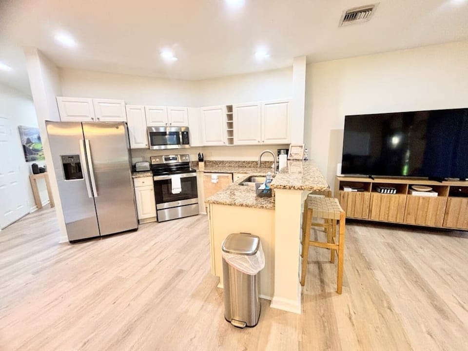 Modern open-concept kitchen and living area featuring stainless steel appliances, granite countertops, breakfast bar seating, wood-look flooring, and a large TV for relaxing or entertaining.