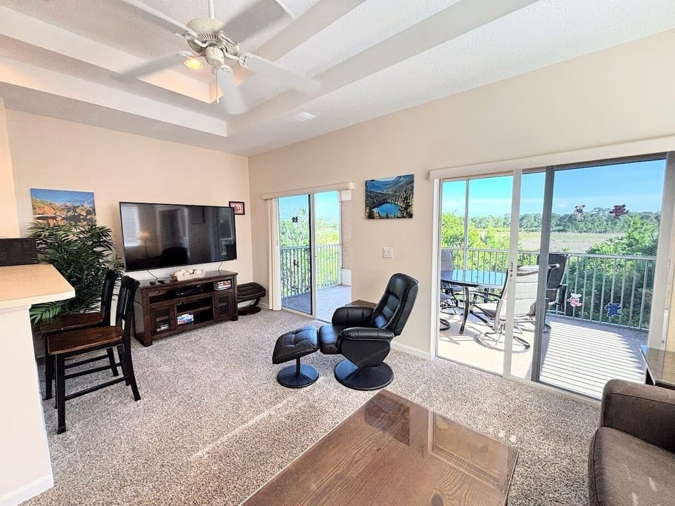 Spacious living area with large TV, cozy seating, and sliding glass doors leading to the screened balcony with peaceful views.