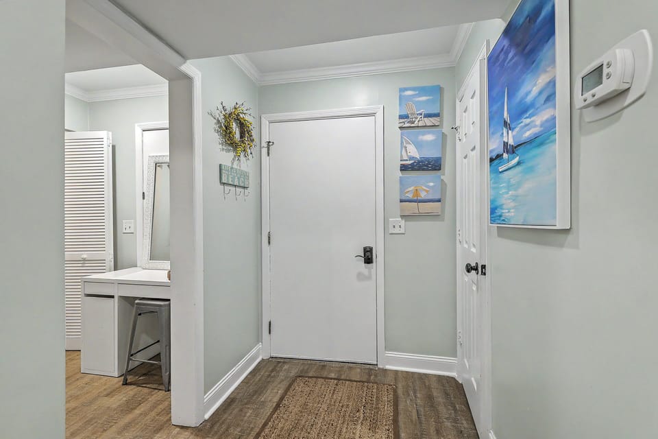 The main entryway foyer features a crisp white door, durable wood-look flooring, and a natural fiber mat. Coastal sailboat artwork and a decorative beach sign add a warm welcome to the cottage.