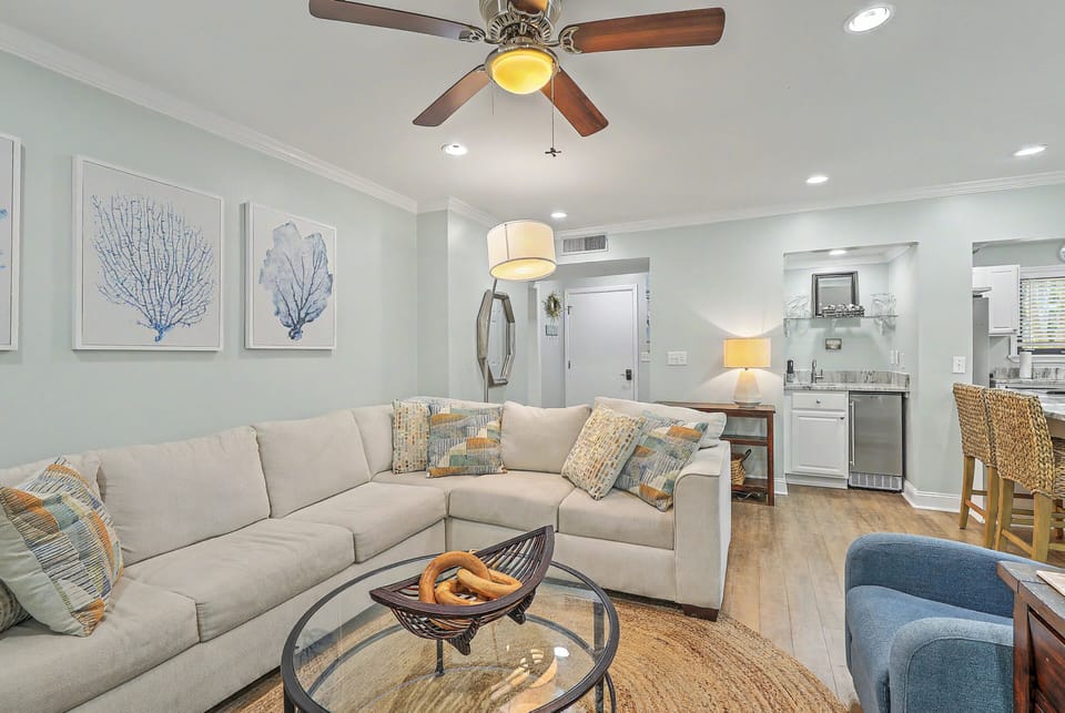A spacious sectional sofa with beach-themed accent pillows provides ample seating in the open living room. A glass coffee table sits on a round jute rug, while a mini-fridge wet bar station serves the space in the background.