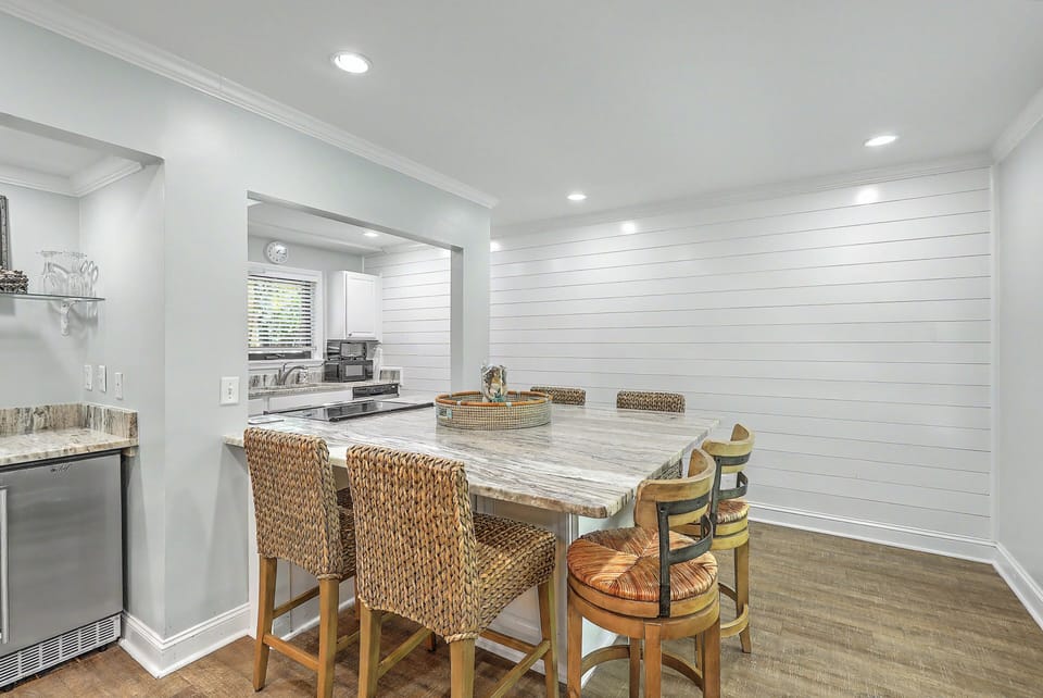 An alternate angle of the central island showcases the modern coastal dining area. Textured barstools line the stone-patterned surface against a crisp white shiplap accent wall with a built-in beverage station.