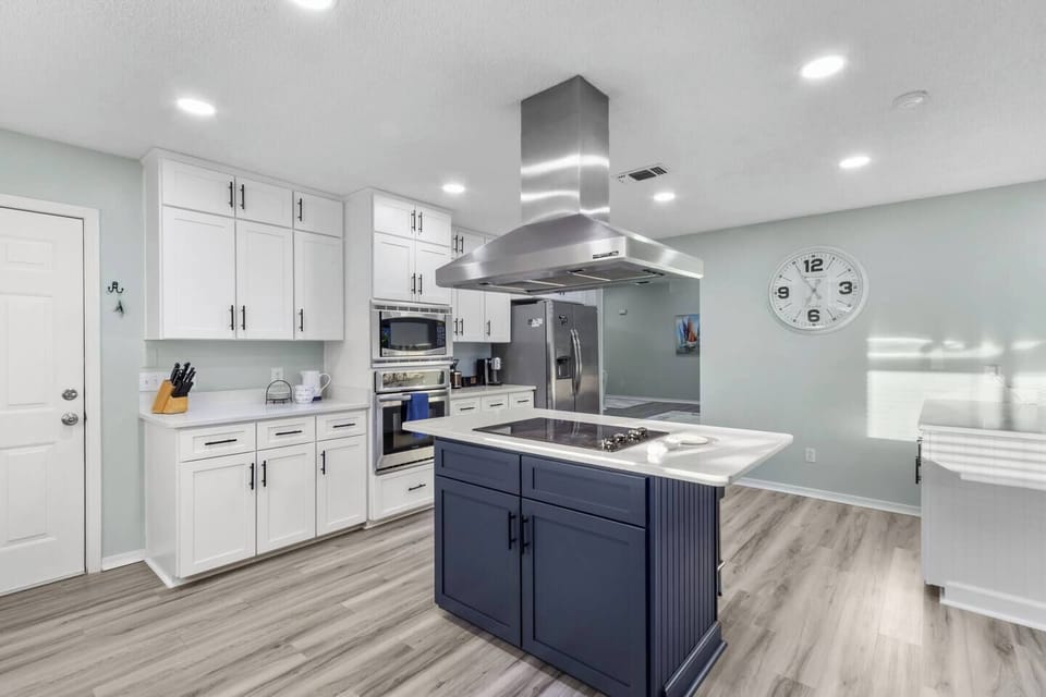 Sleek coastal kitchen with a bold navy island, white quartz countertop, stainless steel island hood, and white shaker cabinets. Premium stainless appliances, light hardwood floors, and a large wall clock complete this stylish, functional space.
