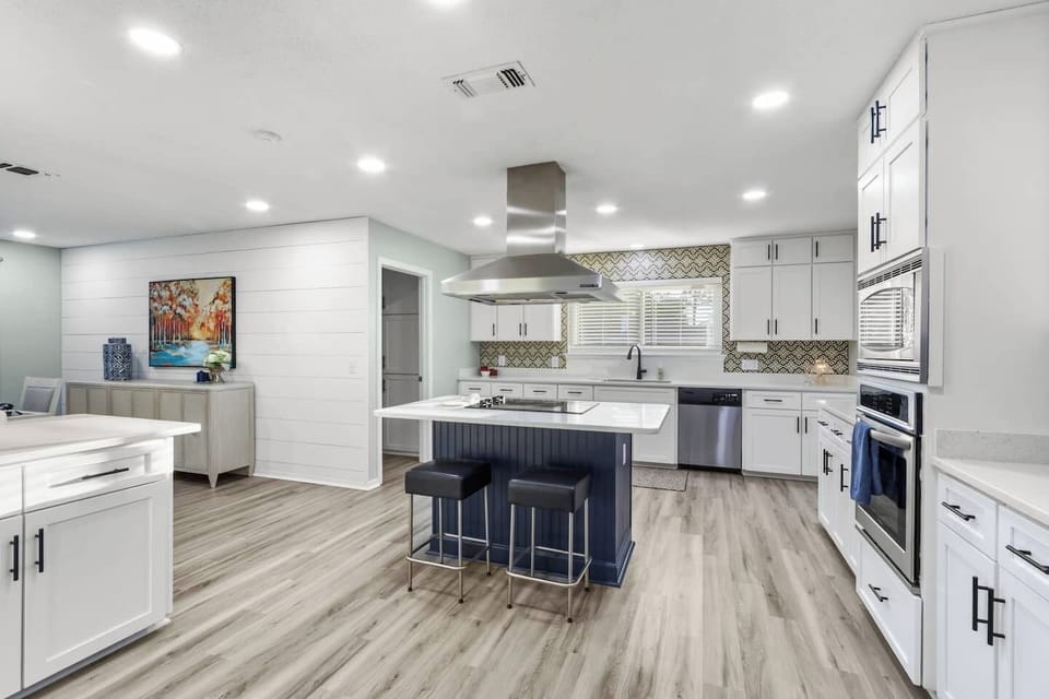 Spacious kitchen with a navy island, black barstools, stainless steel hood, white shaker cabinets, and a bold geometric tile backsplash. Light hardwood floors and a white shiplap accent wall connect seamlessly to the dining area beyond.