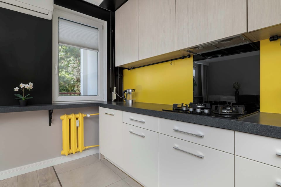 A functional corner of the kitchen reveals a window looking out onto green trees, letting in pleasant daylight. A bright yellow radiator under the counter coordinates perfectly with the vibrant kitchen backsplash.