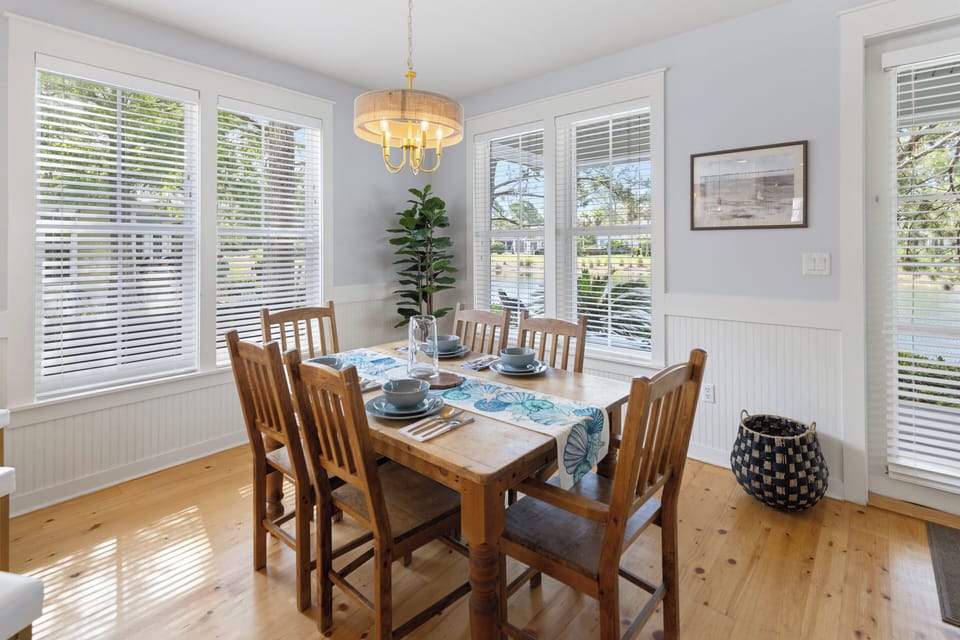 Gather around this sunlit dining table for memorable meals together, with warm wood tones, coastal accents, and bright windows framing leafy garden views that bring the outdoors in.