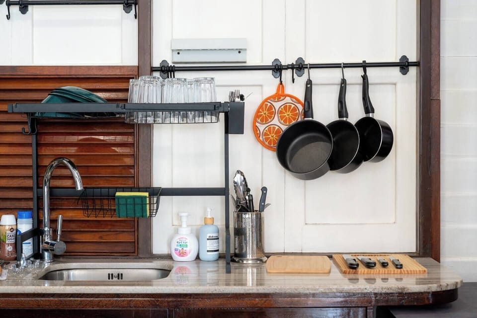 Compact kitchen setup with a sink and faucet, dish rack holding plates and glasses, hanging pans and a citrus-patterned pot holder, utensils in a metal holder, soap dispensers, and wooden cutting boards with knives neatly arranged.