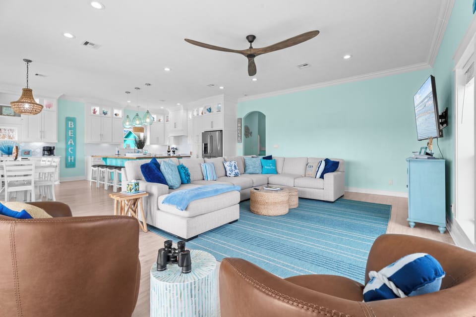 Open-concept living room with sectional sofa, blue area rug, ceiling fan, coastal decor and kitchen island in the background.
