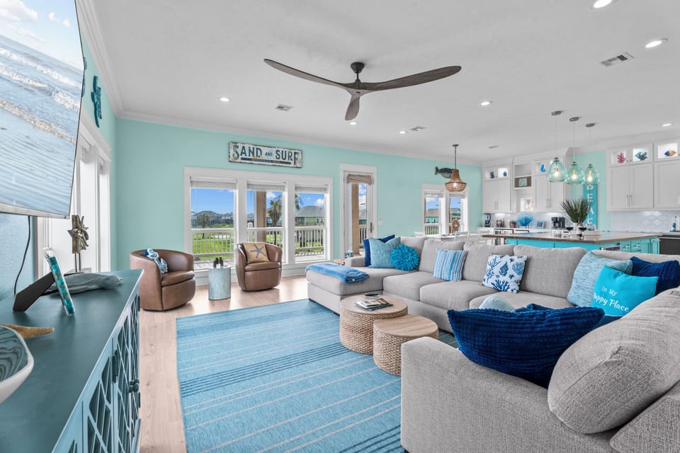 Spacious living area with sectional sofa, lounge chairs, blue rug, balcony doors, ceiling fan and open kitchen beyond.